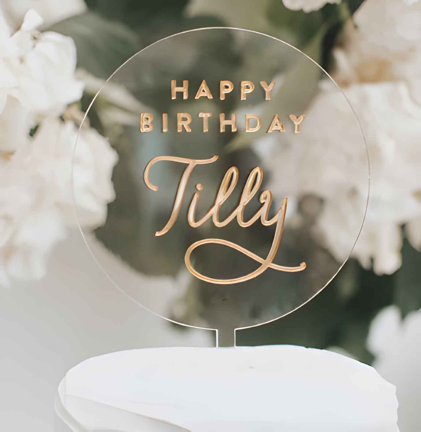 A clear acrylic birthday cake topper features the text "HAPPY BIRTHDAY" in bold, uppercase letters at the top and the name "Tilly" in elegant, cursive script below. The topper is placed on a white cake and is surrounded by soft floral arrangements in the background. The design is modern and celebratory, suitable for a birthday celebration.