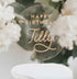 A clear acrylic birthday cake topper features the text "HAPPY BIRTHDAY" in bold, uppercase letters at the top and the name "Tilly" in elegant, cursive script below. The topper is placed on a white cake and is surrounded by soft floral arrangements in the background. The design is modern and celebratory, suitable for a birthday celebration.