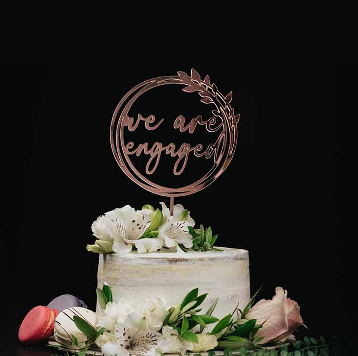 A cake topped with a rose gold acrylic topper that reads "we are engaged" inside a circular frame. The cake has a rustic, textured appearance and is adorned with fresh white flowers and greenery. To the side of the cake, there are colorful macarons, including a pink one. The background is black, highlighting the cake and its decorations.