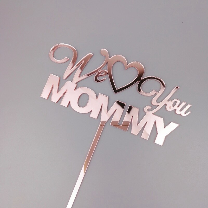 An acrylic cake topper featuring the words "We Love You Mommy" in a decorative, cursive font. The topper is designed in a shiny rose gold color and includes a heart symbol between "Love" and "You." The words are arranged with "We" and "You" in a flowing script, while "LOVE" is prominently featured in a bolder font. The topper has a long stick for easy insertion into a cake. The background is a neutral gray color.
