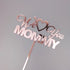 An acrylic cake topper featuring the words "We Love You Mommy" in a decorative, cursive font. The topper is designed in a shiny rose gold color and includes a heart symbol between "Love" and "You." The words are arranged with "We" and "You" in a flowing script, while "LOVE" is prominently featured in a bolder font. The topper has a long stick for easy insertion into a cake. The background is a neutral gray color.