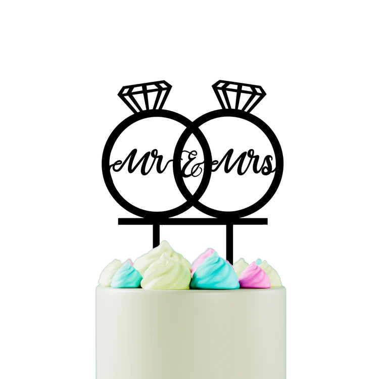 A black cake topper featuring two interlocking wedding rings with the words "Mr" and "Mrs" in elegant script. The topper is placed on a frosted cake with pastel-colored swirl decorations on top, including shades of pink, blue, and green.