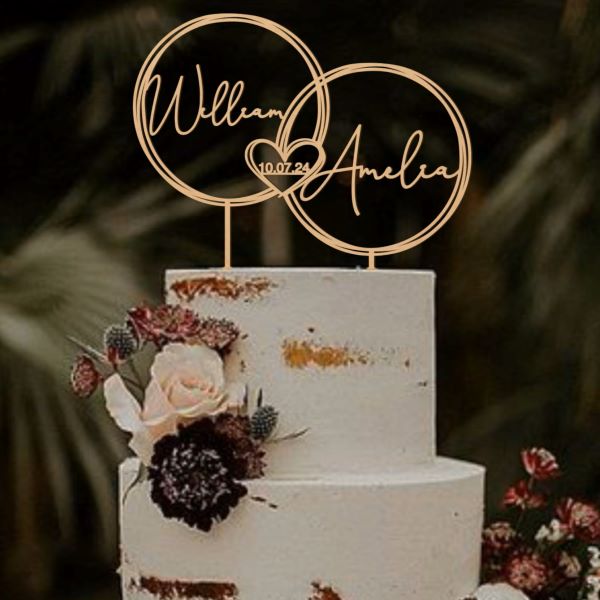 A three-tiered white wedding cake with a textured finish, decorated with hints of gold. Atop the cake is a personalised acrylic topper featuring two interlocking circles. One circle has the name "William" and the other has the name "Amelia" along with the date "10.07.24" inside a heart. The cake is adorned with fresh flowers, including white and dark-colored blooms, enhancing its elegant appearance against a blurred green background.