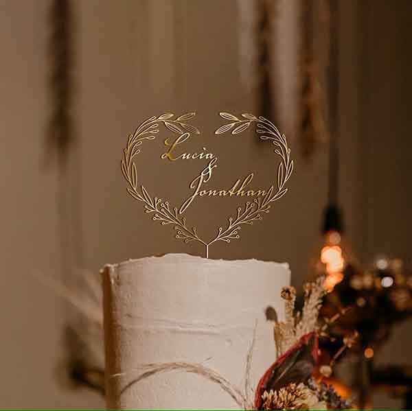 A personalized gold acrylic cake topper in the shape of a heart is displayed on top of a white cake. The topper features foliage designs with the names "Lucia" and "Jonathan" elegantly inscribed inside the heart. The background suggests a festive setting with soft lighting and decorative elements.