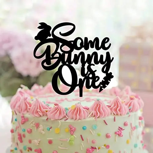 A pastel-colored cake topped with pink frosting, colorful sprinkles, and a black acrylic cake topper that reads "Some Bunny is One." The topper features a silhouette of a bunny and a carrot.