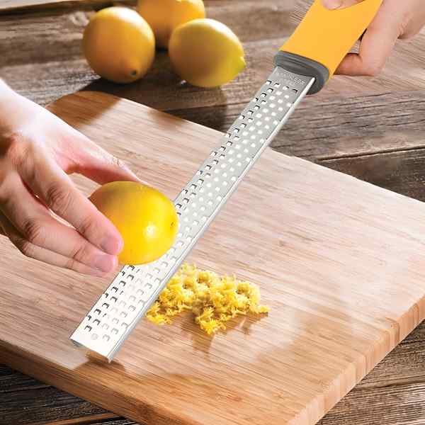A stainless steel zester being used to zest a lemon on a wooden cutting board, surrounded by fresh lemons, highlighting its precision and ease of use.