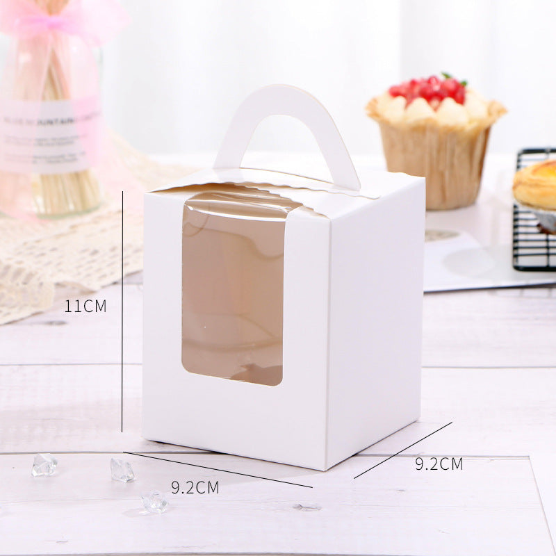 A white cupcake box designed for single portions, featuring a clear front window. The box dimensions are approximately 11 cm in height and 9.2 cm in width and depth. It has a sturdy handle on top for easy carrying. In the background, there are baked goods visible, including a decorated cupcake and possibly other pastries on a wire rack. Small decorative crystals are scattered on the surface around the box.