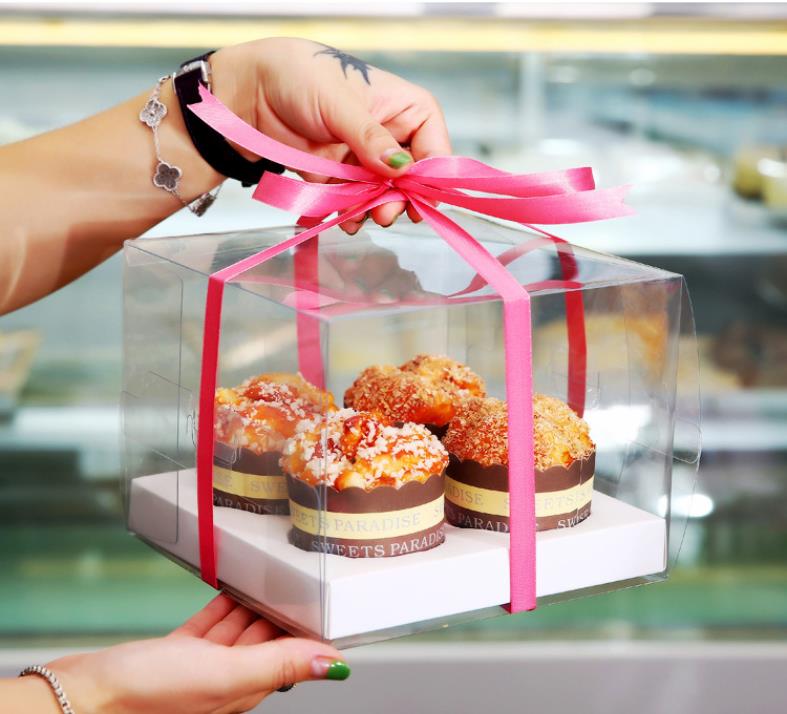 A hand holding a clear cupcake box tied with a pink ribbon, containing four decorated cupcakes arranged on a white base. The cupcakes feature various toppings and are presented in brown cupcake liners with a decorative label. In the background, a display case with more baked goods is visible.