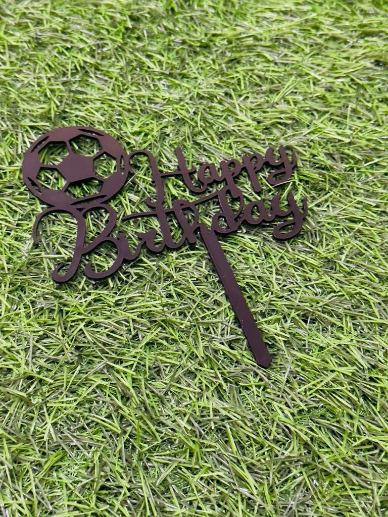 A cake topper featuring the phrase &quot;Happy Birthday&quot; in an elegant script. It is designed with a soccer ball at the beginning of the text. The topper is placed on a green grass-like background, emphasizing its decorative purpose for a birthday celebration.