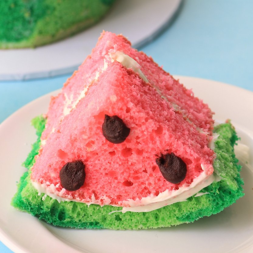 A slice of cake shaped like a watermelon, featuring pink sponge cake for the flesh, accented with green sponge cake for the rind. The slice has three dark chocolate pieces resembling watermelon seeds and is adorned with a layer of white frosting. The background is a light blue.