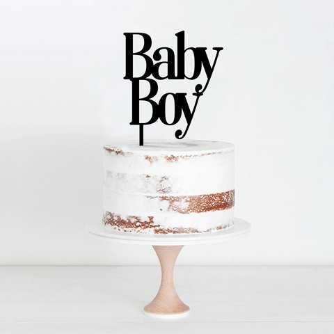 An unfinished, two-layer white cake with a rustic naked design, displaying hints of brown in the frosting, sits on a wooden cake stand. Topping the cake is a black acrylic cake topper that reads &quot;Baby Boy&quot; in playful typography. The background is light and neutral, enhancing the focus on the cake.