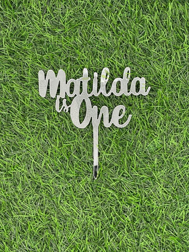 A decorative acrylic cake topper featuring the text &quot;Matilda is One&quot; in a stylish script font. The topper has a stake for easy insertion into a cake. It is displayed against a background that resembles green grass or artificial turf.