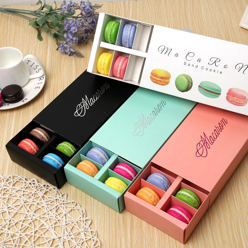 A display of colorful macaron boxes containing various flavored macarons. There are three boxes: one black, one light blue, and one peach. Each box is partially open, showcasing four macarons in different colors such as pink, green, yellow, and orange. The top of the image features a white box with additional macarons, including shades of blue and purple. A small white cup and a black macaron are visible on a round plate in the background, along with some decorative flowers.