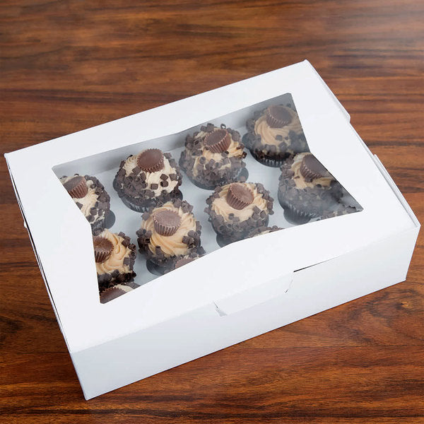 A white cupcake box with a clear plastic window showing nine chocolate cupcakes decorated with swirls of frosting, chocolate chips, and miniature chocolate cups on top. The box is placed on a wooden surface.