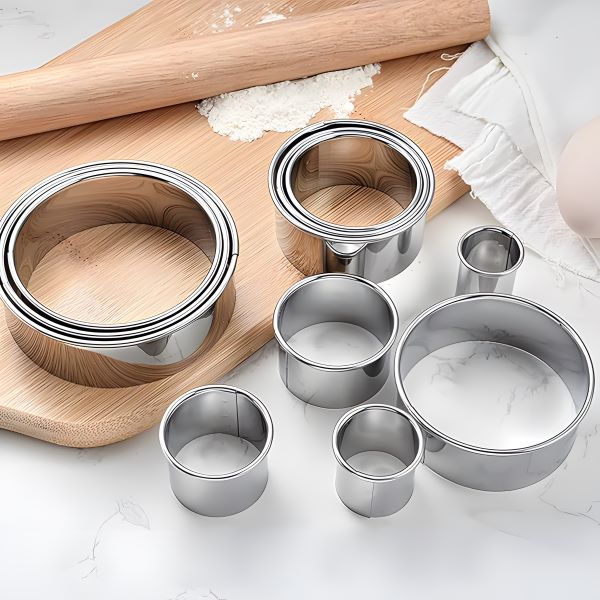 A set of 12 stainless steel round cookie cutters placed on a wooden cutting board, surrounded by baking tools like a rolling pin, flour, and a kitchen towel. The cutters vary in size, highlighting their versatility for baking and food preparation.