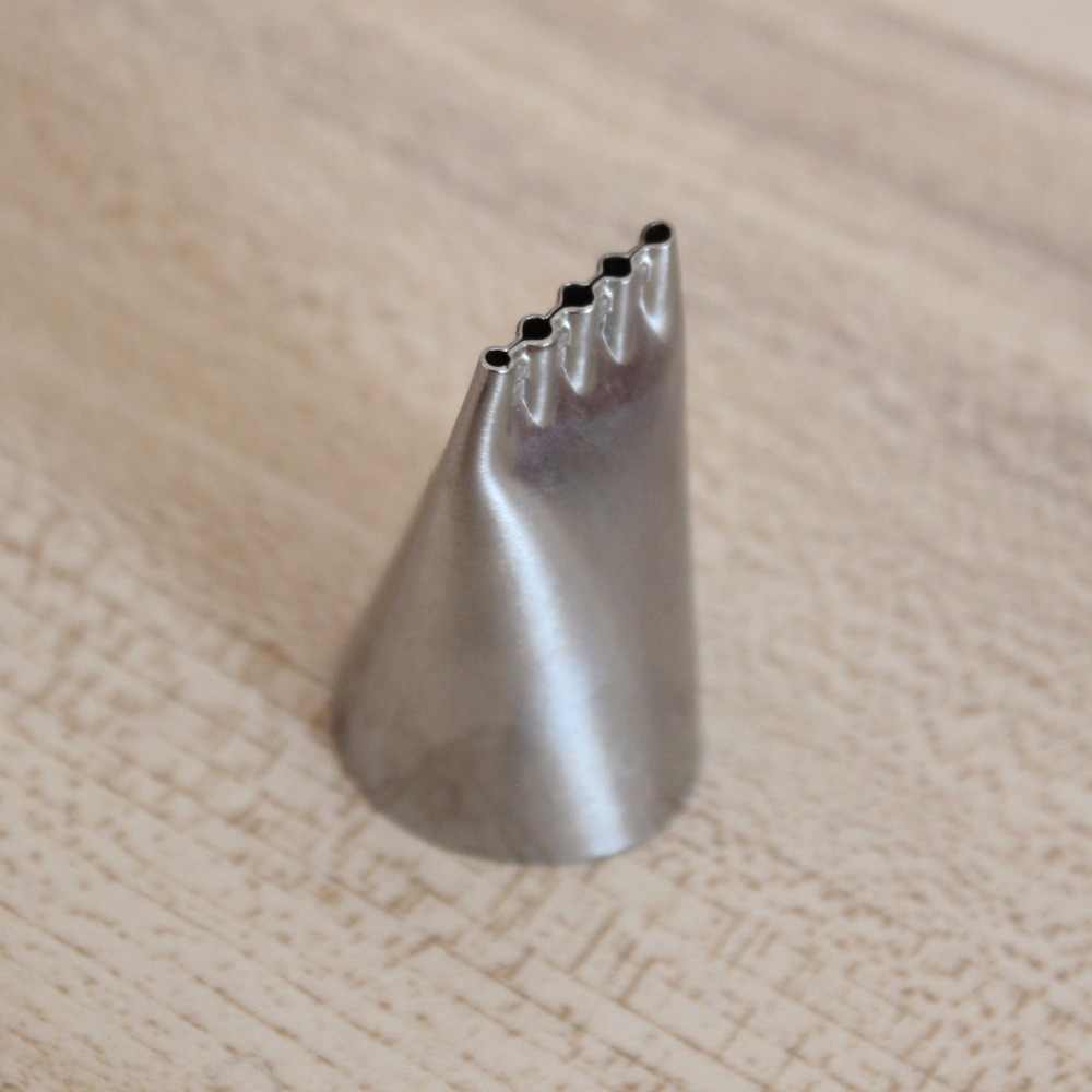 A stainless steel piping nozzle is shown standing upright on a wooden surface. The nozzle has a conical shape with a fluted opening at the top, featuring five rounded tips. Its smooth surface reflects light, highlighting its metallic finish. The background is a light wood grain texture.