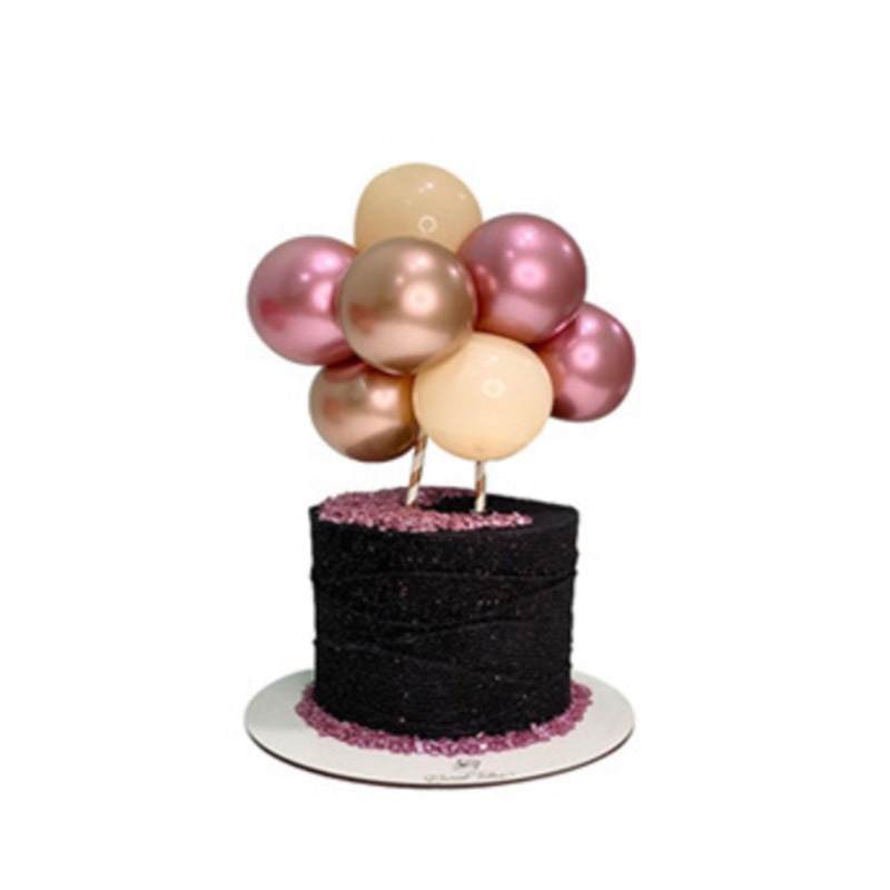 A cake decorated with a cluster of balloons is displayed as a cake topper. The cake is round, covered in black material with a glittery texture and is topped with a layer of pink confetti. The balloon cluster consists of ten balloons in various shades, including light pink, dark pink, and beige, which are attached to two thin sticks. The cake sits on a round white pedestal, enhancing its presentation.