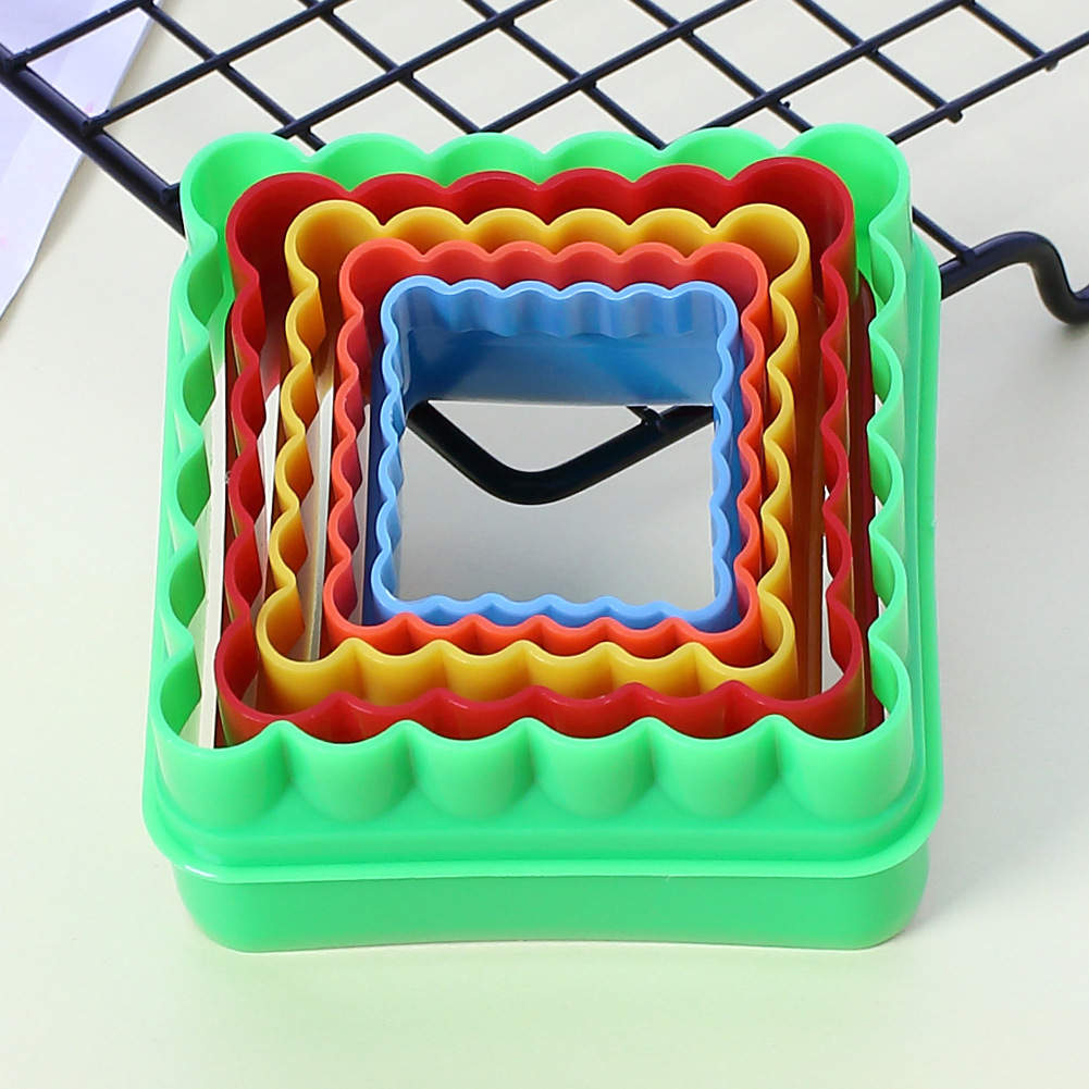 A set of five square cookie cutters is displayed, featuring a variety of bright colors including green, red, yellow, blue, and orange. The cutters are nested together, with the smallest blue cutter positioned in the center and the larger ones arranged around it. The edges of the cutters are scalloped, adding a decorative touch. The background includes a wire rack and a light-colored surface.