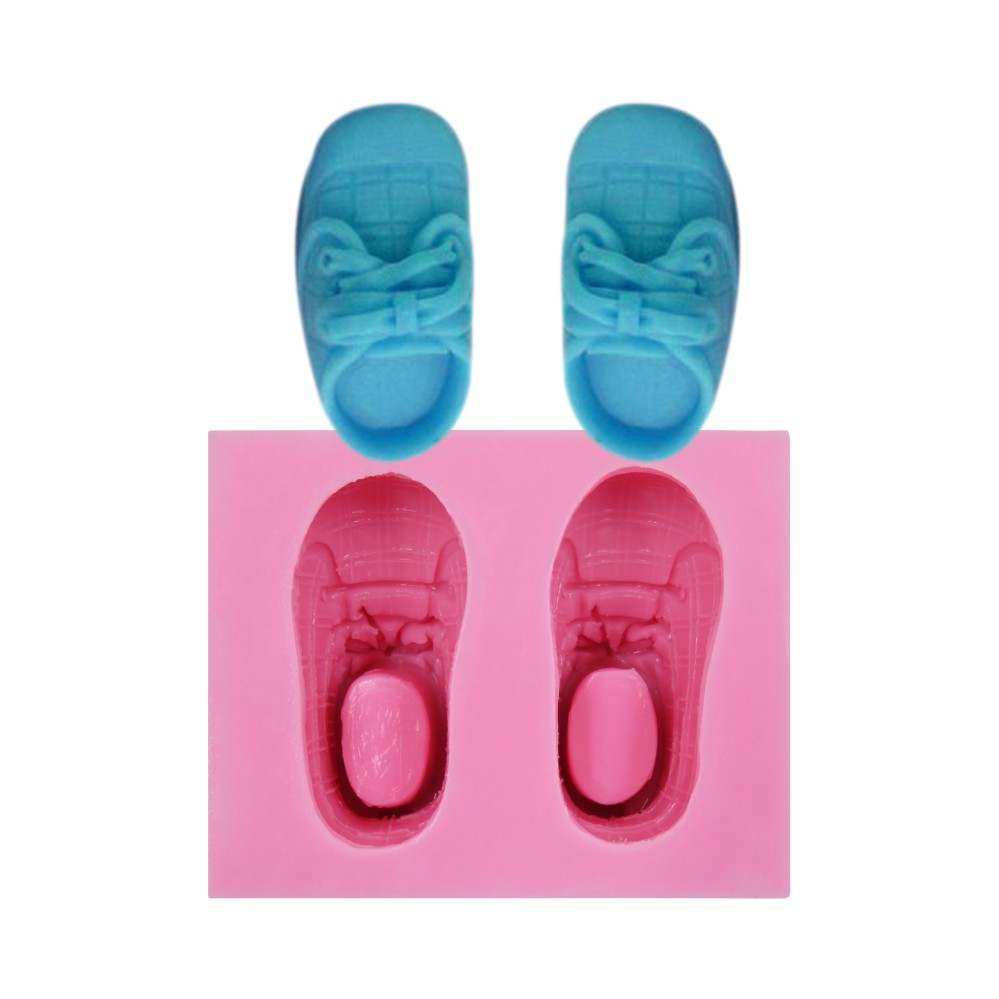 The image shows a silicone mold for baby shoes, with two completed blue baby shoe shapes positioned above a pink mold. The pink mold features detailed impressions of the baby shoes, including laces and sole patterns. The mold and shoes are displayed on a plain background.