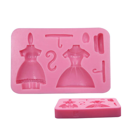 A pink silicone mold designed for creating miniature girl dress and hanger shapes. The main mold features two dress designs, a hanger, a hook, and a small round accessory. A smaller, separate molded piece showing one dress shape is positioned in front of the main mold.