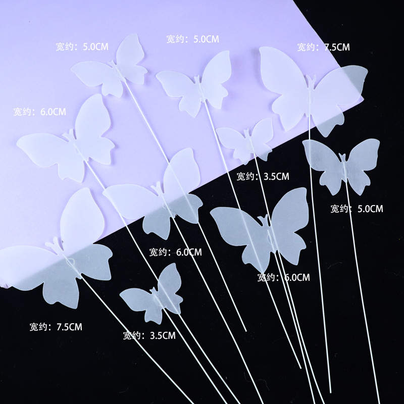 An arrangement of ten white butterfly cake toppers, each secured on a thin stick. The butterflies come in various sizes, with measurements labeled in centimeters next to each one. The background includes both a black and a light purple surface, highlighting the transparent, delicate appearance of the toppers.