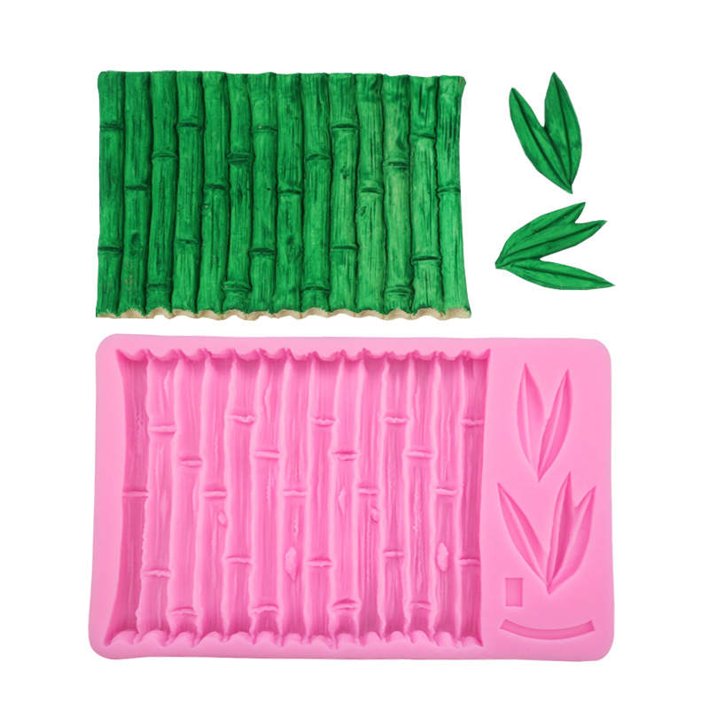 A pink silicone mould featuring a bamboo design is placed below a small, green, textured piece resembling bamboo, along with several green leaves. The mould has grooves that replicate the look of bamboo stalks and includes leaf patterns on one side.