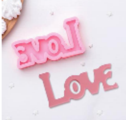 The image shows two silicone molds shaped like the word "LOVE." One mold is in a light pink shade and features the word in uppercase and lowercase letters, with the letters connected in a cursive style. The other mold is a darker pink and also displays the word "Love" in a block lettering style. Both molds are placed against a white background that includes small star-shaped decorations.
