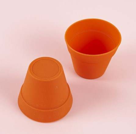 Two orange silicone plant pot molds are shown on a light pink background. One mold is inverted, displaying its base, while the other is right side up, revealing its interior. Both molds have a tapered design typical of plant pots.