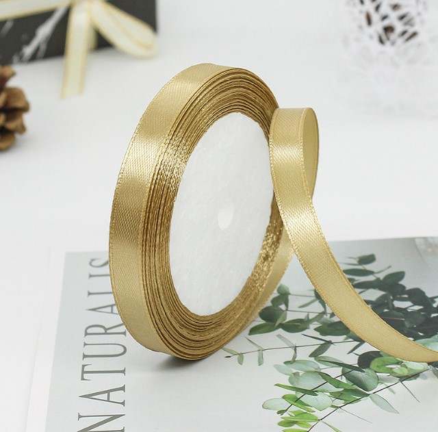 A spool of gold ribbon, 1 cm wide, is displayed on top of a magazine or booklet with green plant illustrations. The ribbon is partially unwound, showing its smooth, shiny texture against a white circular inner core. The background is slightly blurred, with a gift wrapped in a ribbon visible in the corner.