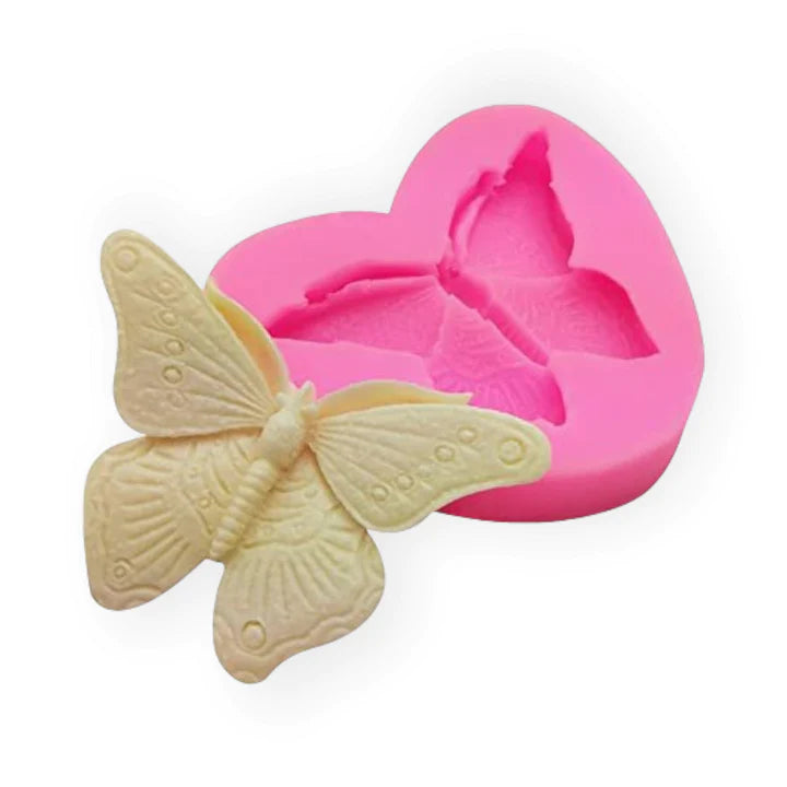 A pink silicone mold shaped like a heart with a butterfly design is displayed next to a white butterfly figure that was made using the mold. The mold features a detailed butterfly imprint, and the finished butterfly figure has textured wings and body.