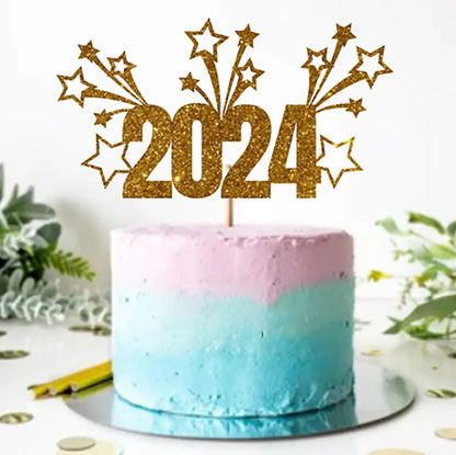 A colorful cake with a gradient design features a light pink top layer and a light blue bottom layer. A gold glittery cake topper displays the year "2024" along with decorative stars and bursts. The cake is placed on a round silver cake board, surrounded by green foliage and scattered gold confetti on the table.