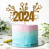 A colorful cake with a gradient design features a light pink top layer and a light blue bottom layer. A gold glittery cake topper displays the year "2024" along with decorative stars and bursts. The cake is placed on a round silver cake board, surrounded by green foliage and scattered gold confetti on the table.