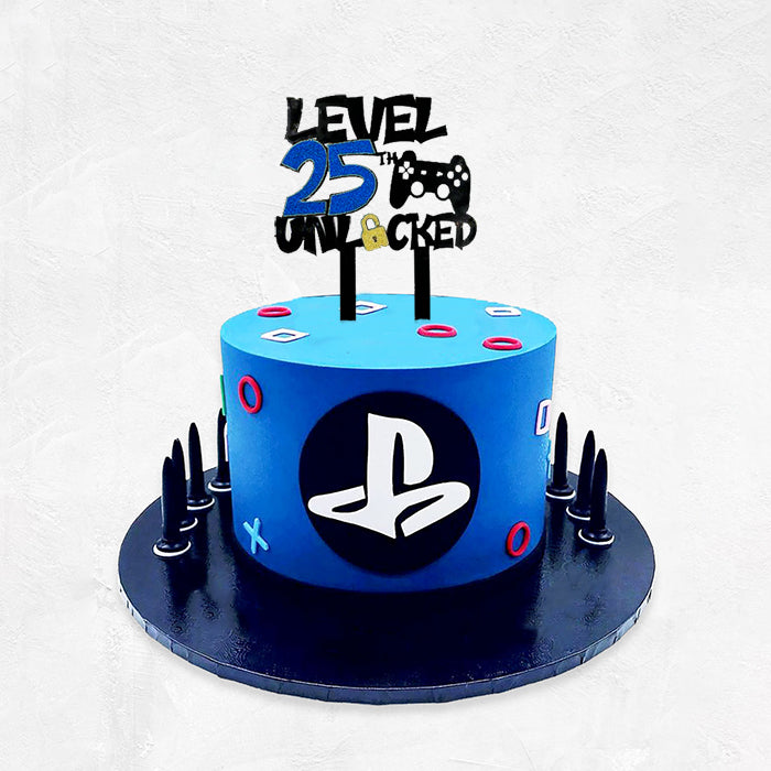 A two-tiered blue cake with a black base. The top is adorned with a black acrylic topper that reads &quot;LEVEL 25th UNLOCKED&quot; in bold letters, alongside a video game controller graphic. The cake features colorful shapes and a PlayStation logo on the front, with small black spikes around the base.