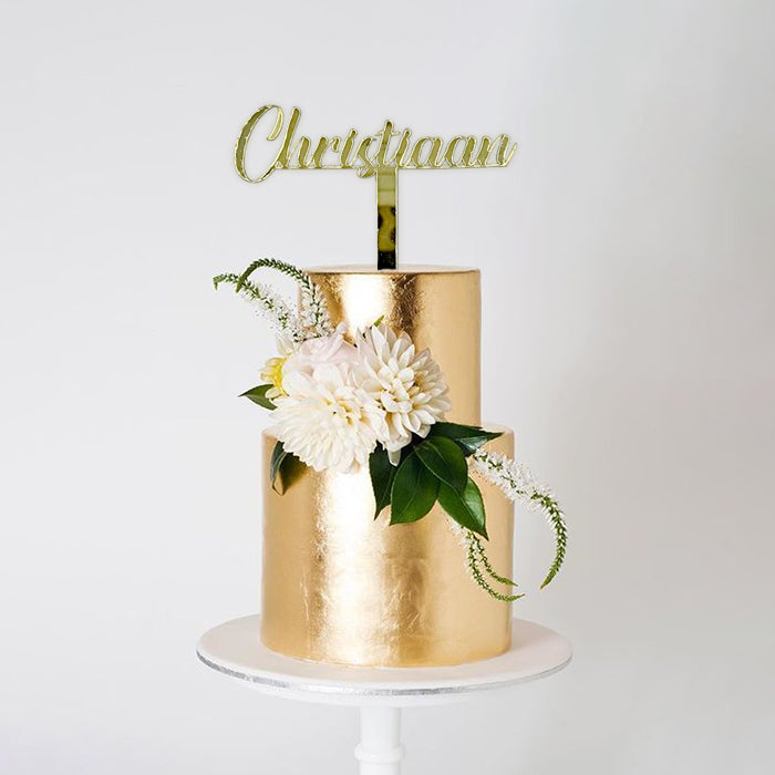 A two-tiered cake with a shiny gold finish, topped with a customized name topper reading &quot;Christiaan.&quot; The cake is adorned with white flowers and green foliage, creating an elegant presentation on a white cake stand against a plain white background.