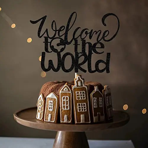 An image depicts a chocolate cake topped with a customized cardstock topper that reads &quot;Welcome to the World&quot; in an elegant, cursive font. The cake is garnished with gingerbread houses around its base, and it is displayed on a wooden cake stand. The background is softly illuminated with string lights, creating a warm atmosphere.