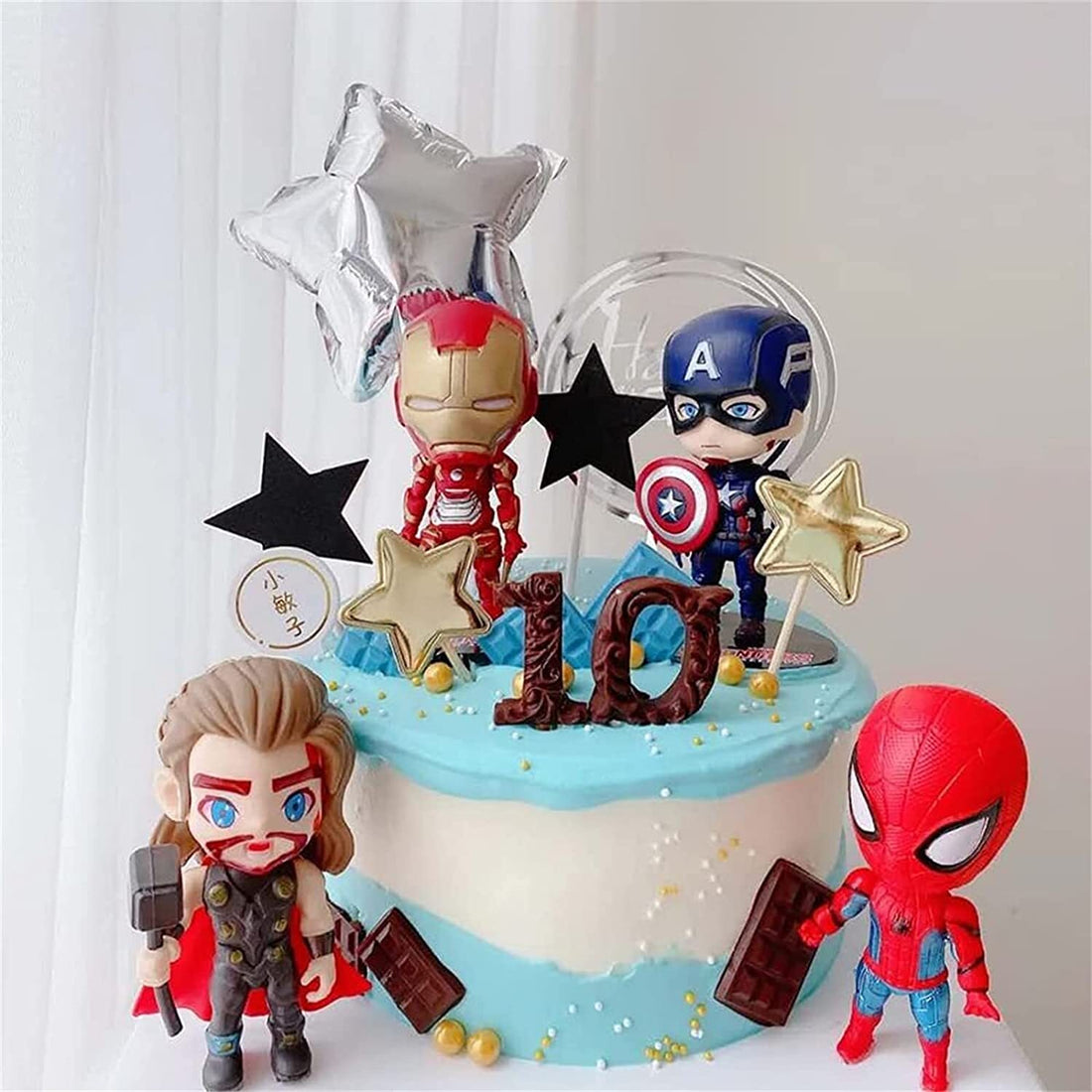 A decorated cake featuring four cartoon superhero cake toppers. The cake is light blue with white frosting and has playful decorations, including stars and a &quot;10&quot; made of chocolate. The toppers include Iron Man, Captain America, Thor, and Spider-Man, each in a fun, animated style. There are also silver star balloons and chocolate bars placed around the cake.