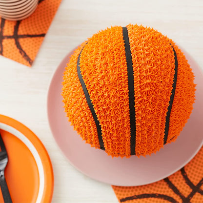 An orange 3D cake shaped like a basketball sits on a pink plate. The cake is detailed with a textured surface resembling basketball dimples and features black icing stripes that mimic the design of an actual basketball. Surrounding the cake are orange-themed party supplies, including a decorative napkin and a colorful plate. The background is a light-colored surface, adding to the festive presentation.