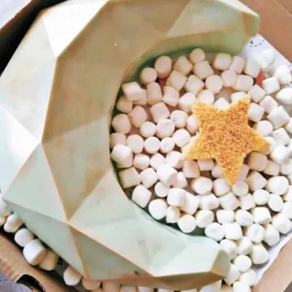 A light green, 3D diamond-shaped silicone mold in the form of a crescent moon, placed in a cardboard box filled with small white packing peanuts. A yellow star-shaped sponge rests in the center of the mold.