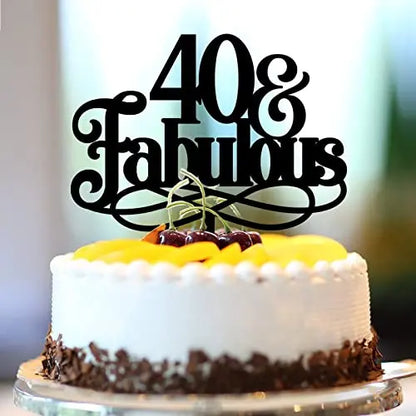 A round cake topped with a black decorative cake topper that reads "40 &amp; Fabulous." The cake is frosted in white with a chocolate border and is garnished with yellow fruit on top, along with a few cherries. The background is softly blurred.