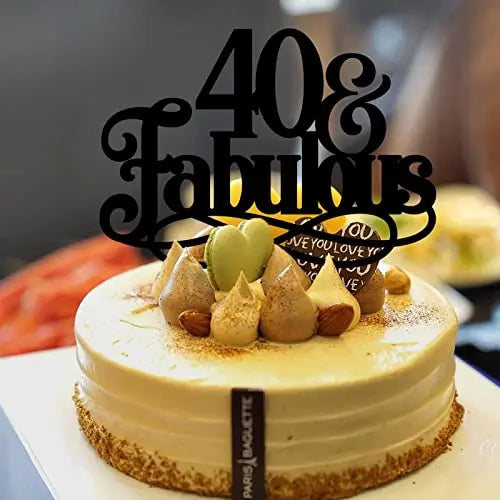 A round cake with a smooth, light-colored frosting is topped with a prominent black cake topper that reads "40 &amp; Fabulous" in an elegant font. The cake is adorned with decorative elements, including small meringues, nuts, and a decorative accent at the base. The background suggests a festive setting.