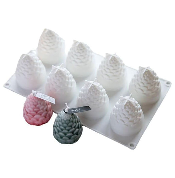 The image features a pine cone silicone mold with ten cavities shaped like pine cones. The mold is white and positioned at the top. In front of the mold, there are three completed candles: two pine cone-shaped candles in pink and green, and several white candles from the mold. Each candle has a wick and a small tag attached. The overall presentation is light and showcases the pine cone design in a clean, bright setting.