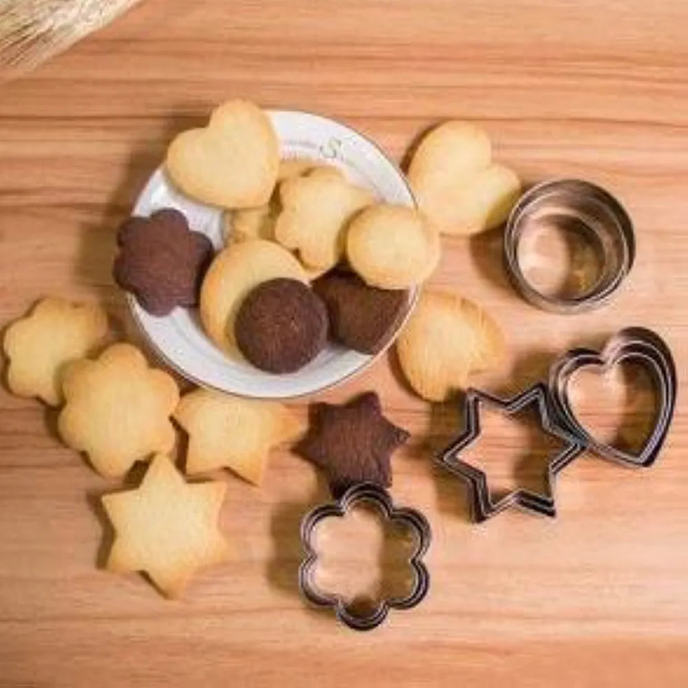 A wooden surface is adorned with various baked cookies, including heart, star, and flower shapes. A small plate at the center holds several cookies, while stainless steel cookie cutters in various shapes are arranged alongside the cookies. The cookies are a mix of light and dark colors, creating a visually appealing display.