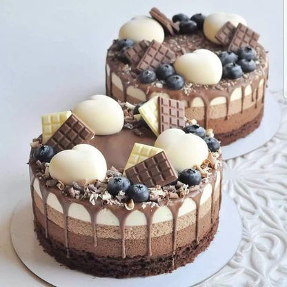 Two round chocolate cakes layered with creamy fillings, topped with chocolate drips, chocolate bars, and white chocolate spheres. The cakes are adorned with fresh blueberries and chocolate shavings, presented on white cake boards against a textured background.