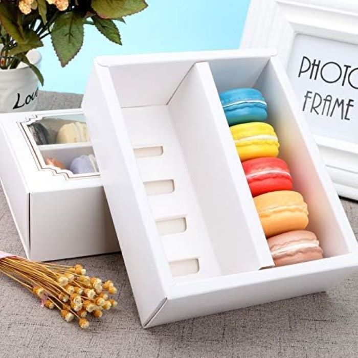 A white macaron box designed to hold 10 portions, partially open to reveal colorful macarons in various hues, including blue, yellow, pink, orange, and red. One side of the box features a small compartment with a transparent window, showcasing additional macarons. The background includes a decorative photo frame and a small bouquet of dried flowers.