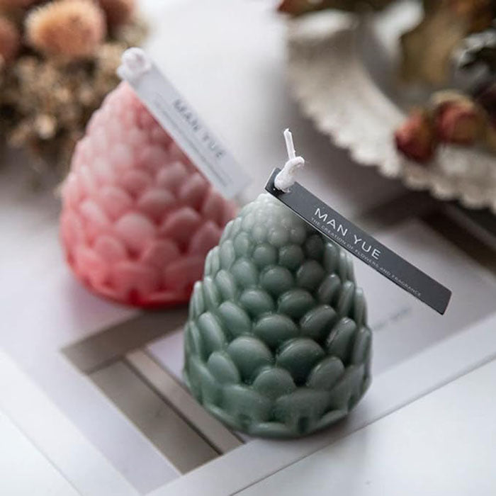 Two decorative candles shaped like pine cones, one in pink and the other in green, are displayed on a light background. Each candle has a wick and a small tag with the brand name "MAN YUE." The arrangement includes soft-focus floral elements in the background.
