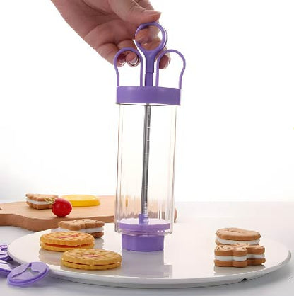 A hand is holding a purple cookie design imprinting tool, which features a clear cylindrical body and a purple base. The tool is placed on a white circular surface surrounded by several colorful cookies, including stacked and decorated varieties. There is also a small red cherry-like decoration and a wooden cutting board in the background.