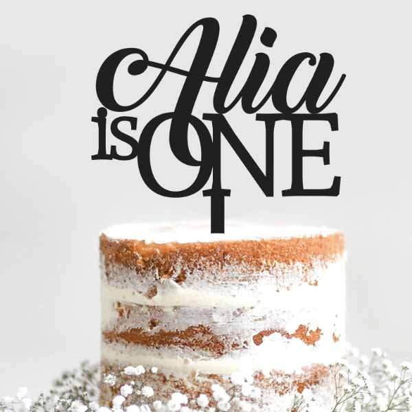 A close-up view of a layered, semi-naked cake topped with a decorative cake topper. The topper features the text &quot;Alia is ONE&quot; in an elegant, modern font. The cake has a rustic appearance with visible layers of cake and frosting, surrounded by small white flowers on the surface. The background is a light gray, enhancing the focus on the cake and topper.