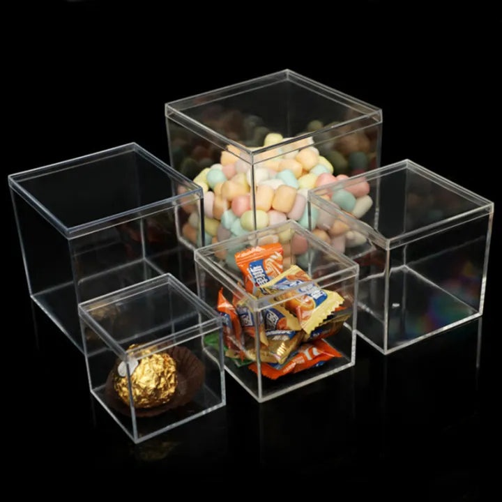 A collection of five transparent acrylic containers of varying sizes. The largest box is filled with colorful, pastel-shaped candy, while a medium-sized box contains small wrapped candies. The smallest boxes are filled with a chocolate truffle and a decorative piece. The containers are arranged on a black surface, creating a contrast that highlights their clarity and the contents inside.