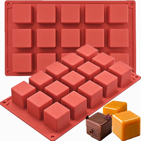 A red silicone mold for making square cubes, featuring a grid of 16 individual cube-shaped cavities. In the foreground, there are two small, colorful cube-shaped chocolates?one yellow and one brown with a decorative ribbon.