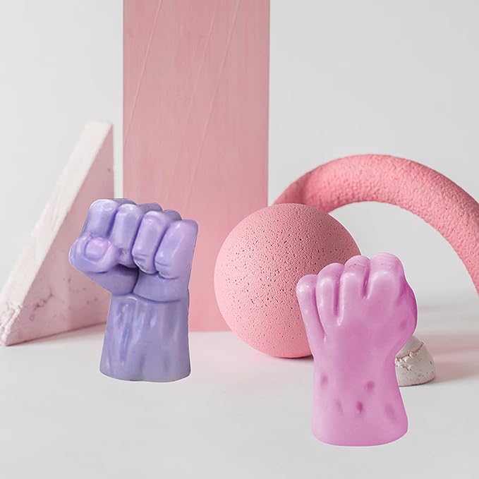 Two colorful silicone fist-shaped molds are prominently displayed in the foreground, one in lavender and the other in pink. The background features a pink vertical block, a round pink object, and a curved pink item, along with a white triangular shape. The arrangement creates a visually appealing composition with soft colors and playful shapes.